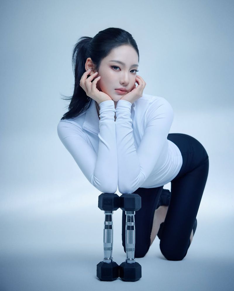 Ningning kneeling and resting her chin in her hands atop two dumbbells in front of a cool-toned studio backdrop.