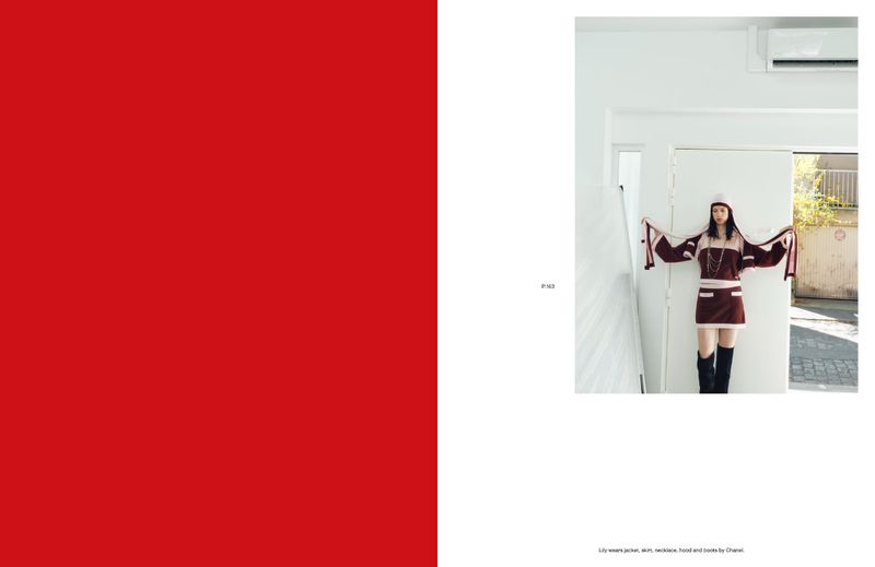 Fashion editorial shot: A young woman in a stylish two-piece outfit and knee-high boots poses in front of a white doorway, brightly lit.