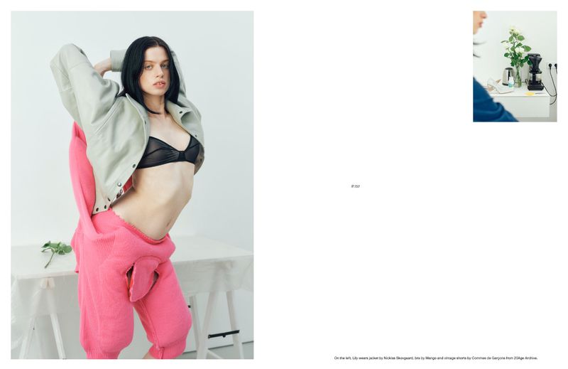 A slender woman with dark hair poses in a black bra, an open pink jumpsuit, and a light grey jacket in a minimalist white studio.