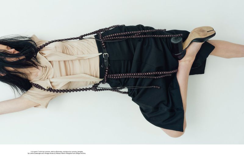 Fashion editorial image featuring a female model in a beige top, black skirt, heels, and beaded strands, lying on a white background.