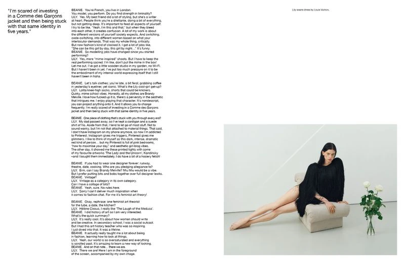 A model lounges on a cream-colored chaise lounge in a minimalist studio, showcasing a dark, fringed fashion garment and white roses.