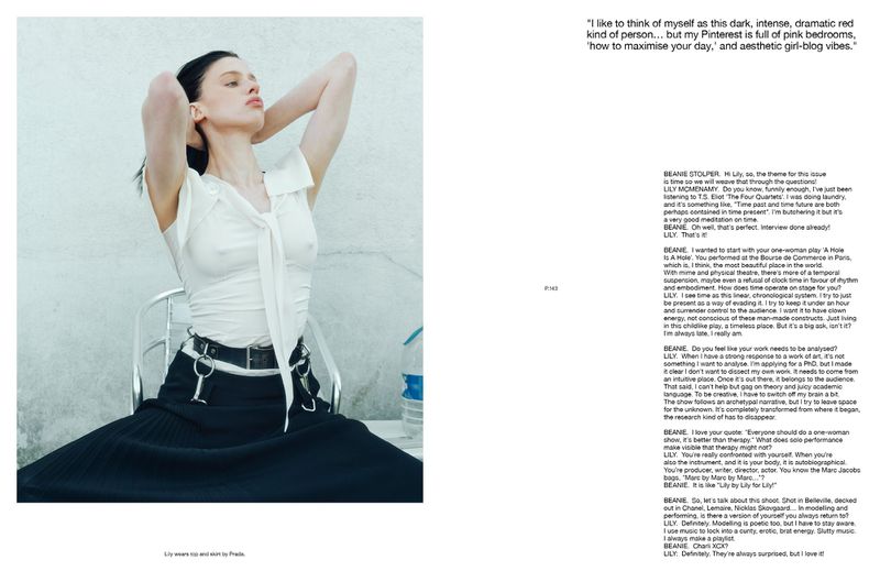 Editorial fashion shot of a woman in a white top and black skirt sitting against a light grey wall in natural light.