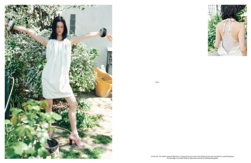 Woman in a white dress poses in a garden setting with flowering bushes, wearing cuff bracelets and pink heeled shoes, arms outstretched.