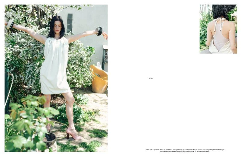 Woman in a white dress poses in a garden setting with flowering bushes, wearing cuff bracelets and pink heeled shoes, arms outstretched.