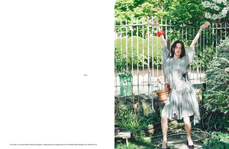A young woman in a pale dress poses outdoors with arms raised in a garden setting, embodying a blend of vintage and contemporary fashion.