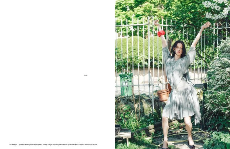 A young woman in a pale dress poses outdoors with arms raised in a garden setting, embodying a blend of vintage and contemporary fashion.