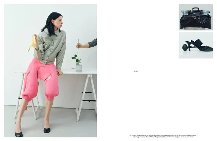Lily McMenamy in a grey jacket and bright pink sculptural shorts seated on a white trestle table holding a banana peel.