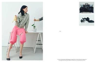 Lily McMenamy in a grey jacket and bright pink sculptural shorts seated on a white trestle table holding a banana peel.