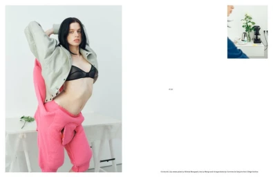 Model Lily McMenamy posing in an oversized grey jacket, sheer black bra, and vibrant pink knit trousers against a white backdrop.