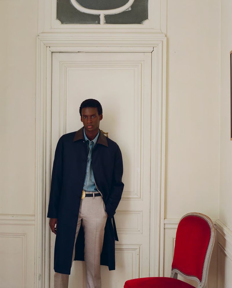 Black male model wearing a navy trench coat and beige trousers standing in front of a white paneled door beside a red chair.