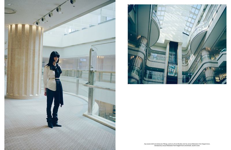 Fashionable woman stands inside a spacious building with geometric architecture; she wears a modern black and white outfit and long black hair.