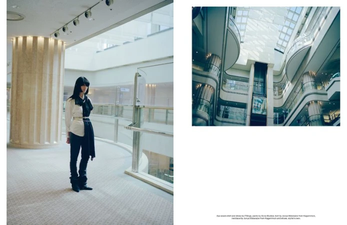 Fashionable woman stands inside a spacious building with geometric architecture; she wears a modern black and white outfit and long black hair.