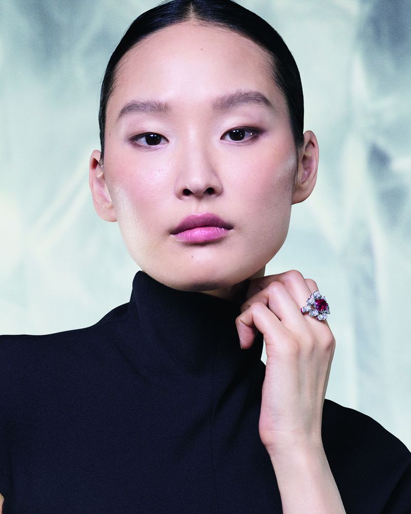 East Asian woman in a black high-neck top showcasing a Cartier ruby and diamond ring against a soft textured background.