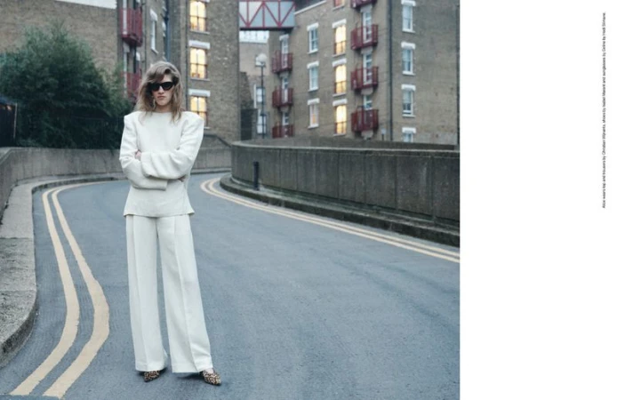 Woman in a white suit with shoulder pads stands on a gray road in front of a brick apartment building with arms crossed.