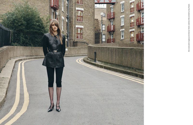 A woman stands on an empty road with yellow lines, wearing a black leather jacket, leggings, and heels, with buildings in the background.