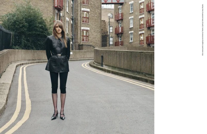 A woman stands on an empty road with yellow lines, wearing a black leather jacket, leggings, and heels, with buildings in the background.