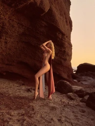 Model Tati K posing in a draped salmon fabric panel against a towering red coastal rock formation at sunset.