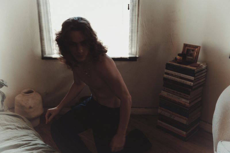 A long-haired, shirtless man is kneeling on the floor in a sparse room with white walls and a window. There's a stack of books.