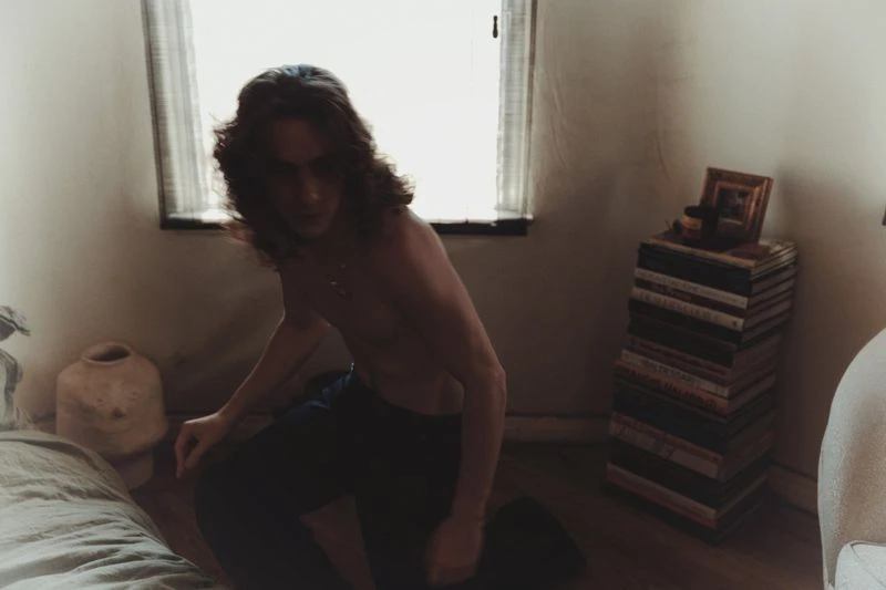 A long-haired, shirtless man is kneeling on the floor in a sparse room with white walls and a window. There's a stack of books.