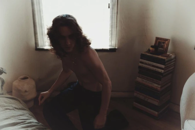 A long-haired, shirtless man is kneeling on the floor in a sparse room with white walls and a window. There's a stack of books.