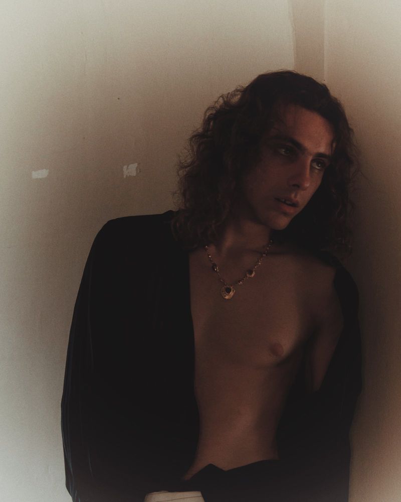 A young adult with long brown hair in a black cardigan stands in front of a white wall, adorned with a necklace, and soft, moody lighting.