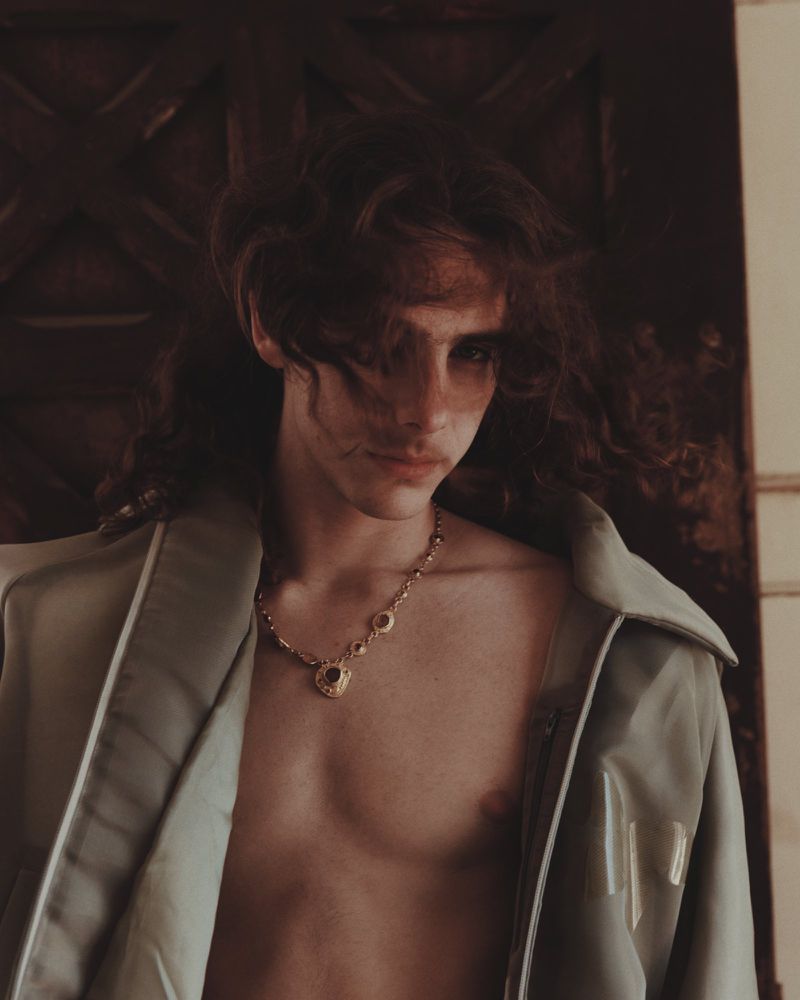 Portrait of a young adult male with long brown hair wearing an unbuttoned, light green jacket and a gold necklace inside a room.