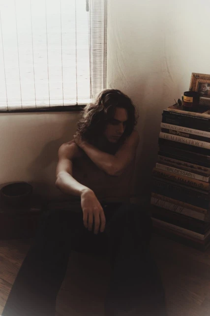 Man with long hair sitting against wall, wearing tank top and pants, next to stack of books, with window blinds behind him.