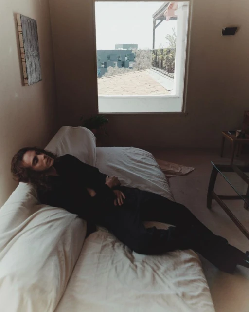 Adult is reclining on a bed in a bedroom interior near a window with bright outdoor light.  Neutral tones and natural light.
