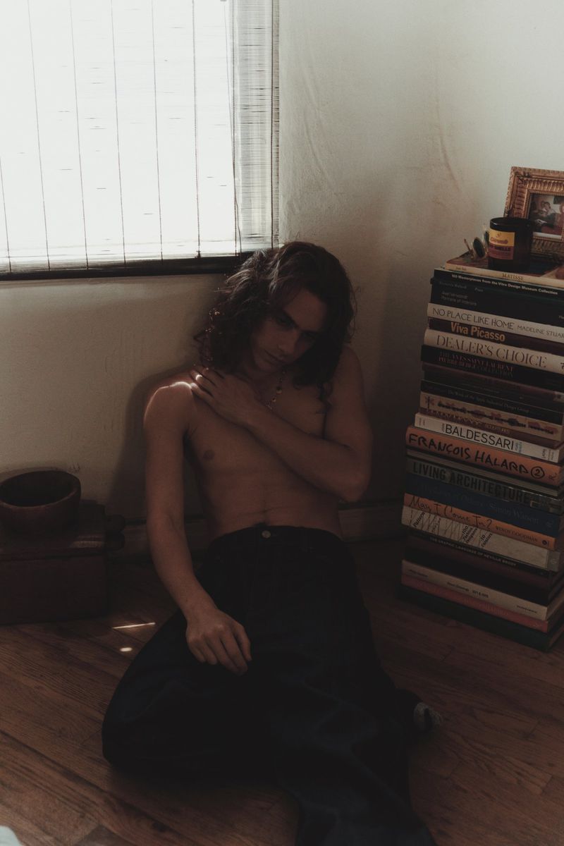 Shirtless young man with wavy hair sitting on a wooden floor beside a tall stack of art books next to a window.
