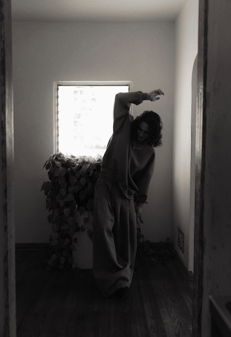 Monochrome portrait of a person in sweatpants and a sweatshirt standing next to a large potted plant in a room with white walls and a window.