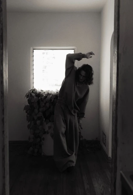 Monochrome portrait of a person in sweatpants and a sweatshirt standing next to a large potted plant in a room with white walls and a window.