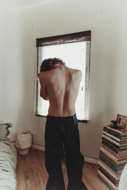 Shirtless person faces a bright window in a sparse bedroom. Stacks of books and personal items suggest domestic interior lifestyle.