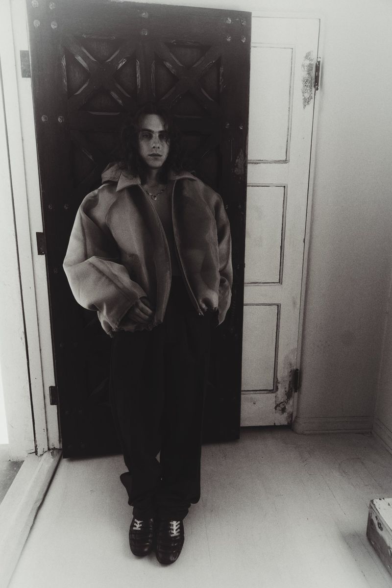 Monochromatic shot of a person with long hair, wearing a jacket and dark pants, standing in front of a partly open door indoors.