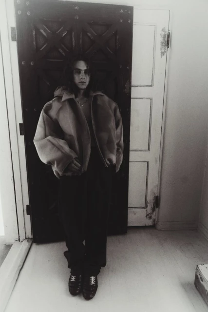 Monochromatic shot of a person with long hair, wearing a jacket and dark pants, standing in front of a partly open door indoors.