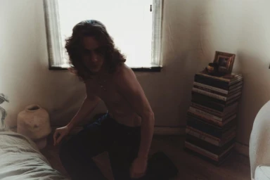 Shirtless man with long dark hair kneeling in a bedroom by a window for Numero Netherlands
