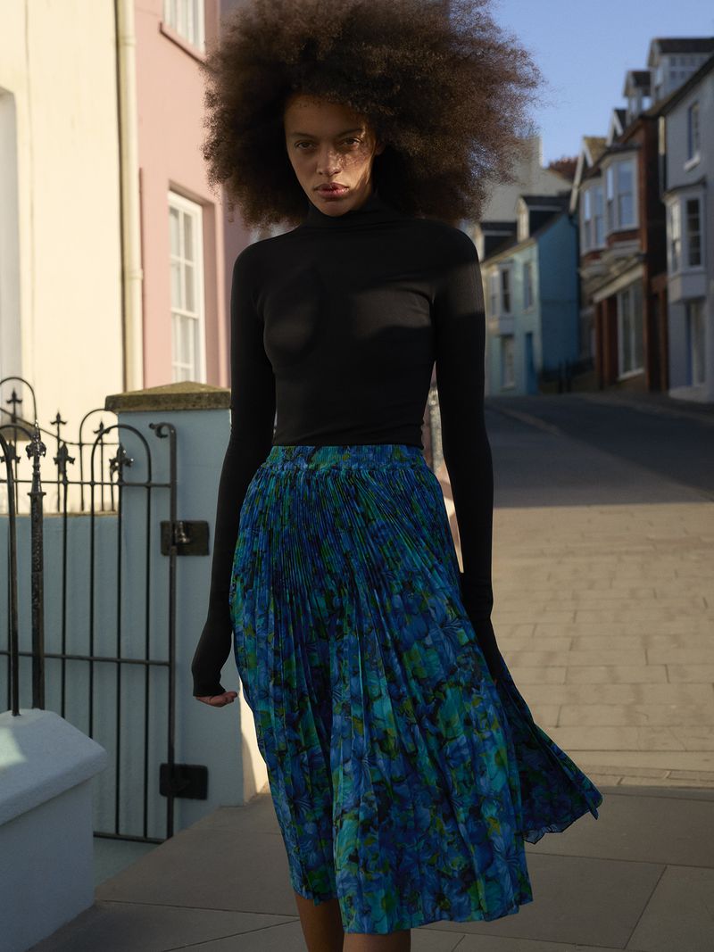 Fashion model with volume afro hair in a black turtleneck and pleated blue skirt standing on a residential street.