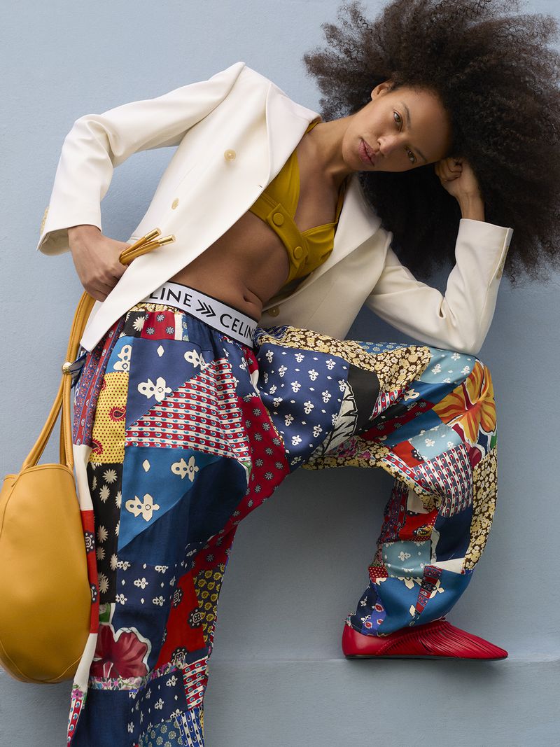Model with voluminous afro hair wearing a white blazer and colourful patchwork trousers leaning against a blue wall.