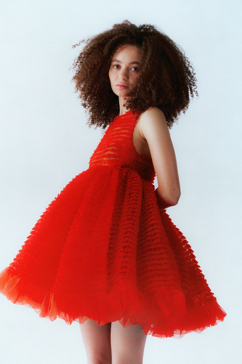 Model with voluminous curly hair wearing a vibrant red ruffled dress against a plain white studio background.