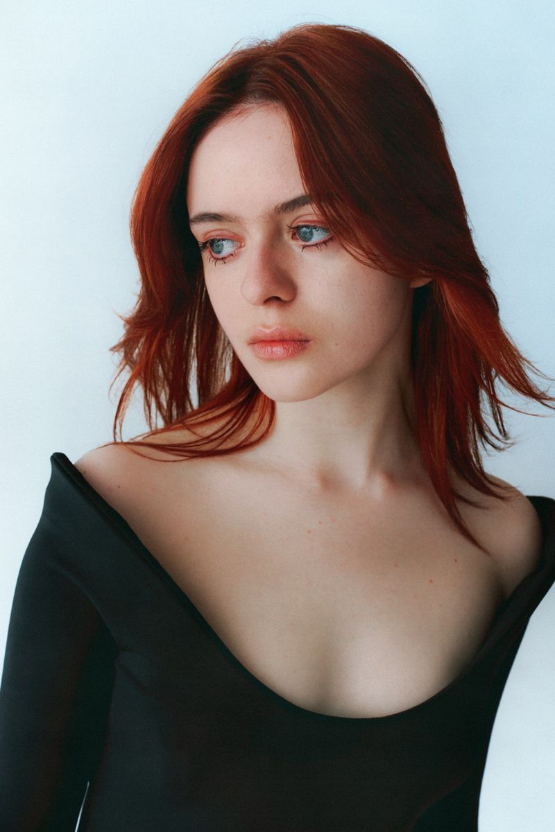Portrait of a young woman with reddish hair wearing a black off-shoulder top against a plain light background