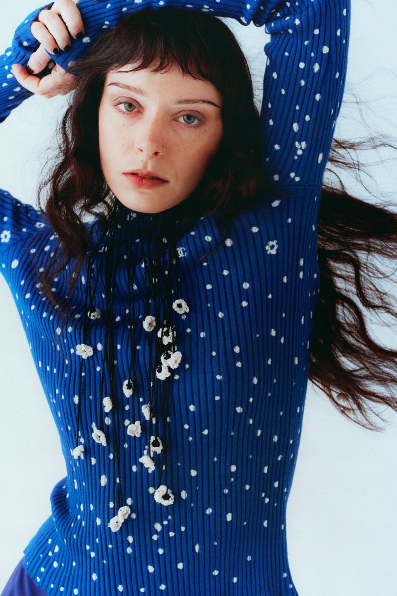 Holly Humberstone wearing a blue speckled ribbed knit turtleneck sweater with hands behind her head against a white backdrop.