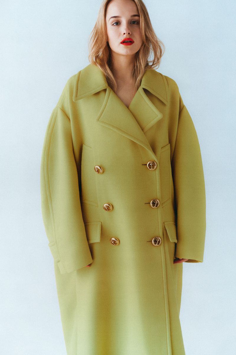 Woman with red lipstick wearing an oversized chartreuse double-breasted wool coat against a pale seamless studio background.
