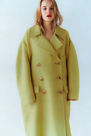 Woman with red lipstick wearing an oversized chartreuse double-breasted wool coat against a pale seamless studio background.