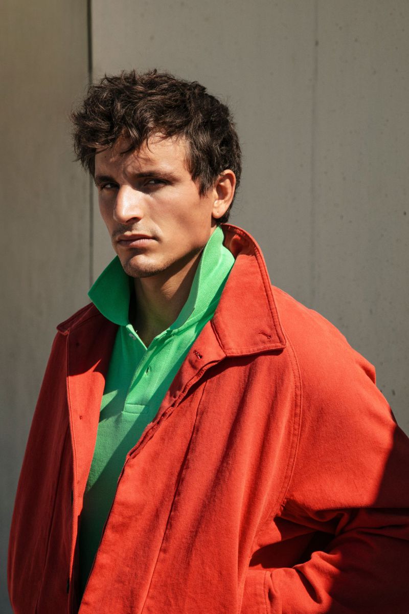 Model Sergio Modrego wearing a bright green polo shirt under a rust-red jacket against a concrete wall in direct sunlight.