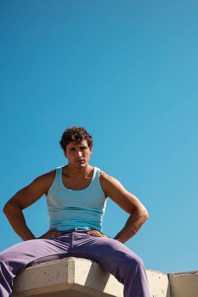 Model Sergio Modrego wearing a light blue tank top and purple pants seated on a concrete ledge against a clear blue sky.