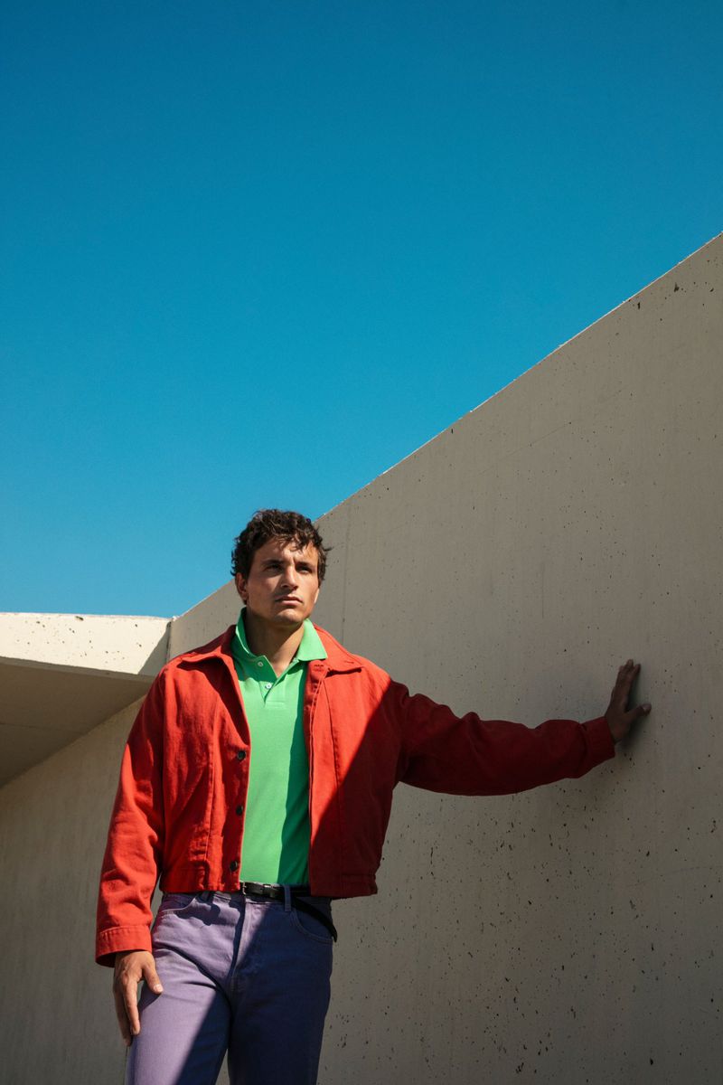 Model Sergio Modrego posing in a red jacket, green polo, and purple trousers against a concrete wall under a clear blue sky.