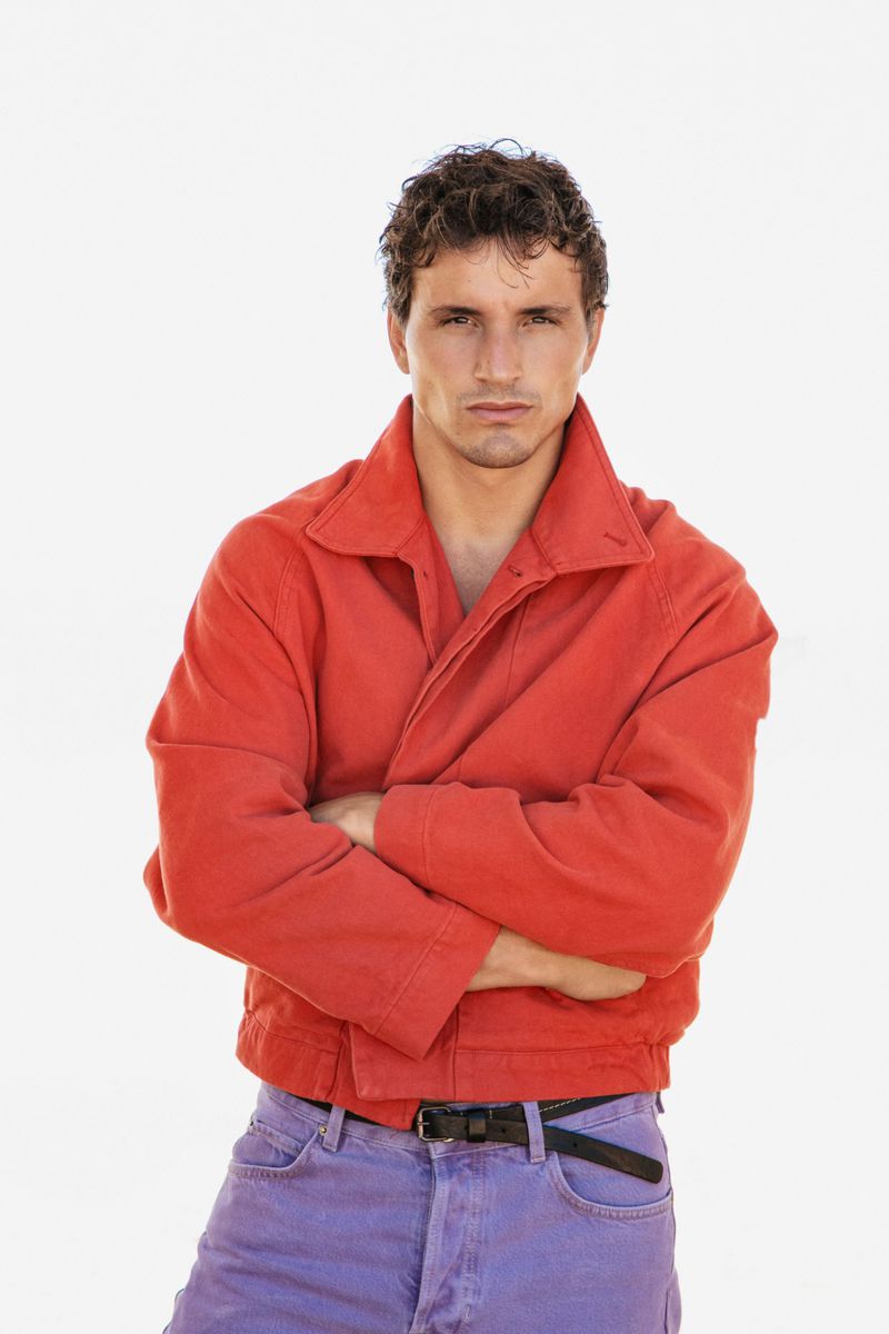 Model Sergio Modrego posing with crossed arms wearing a coral red zip jacket and purple jeans against a white background.