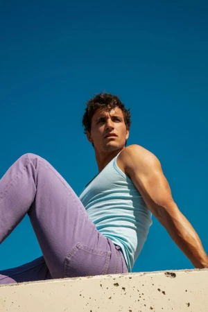 Model Sergio Modrego wearing a light blue tank top and purple denim jeans seated against a clear blue sky.