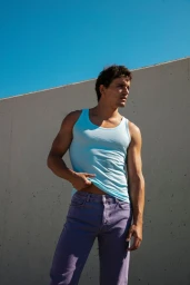 Model Sergio Modrego wearing a light blue tank top and purple denim jeans against a concrete wall under direct sunlight.