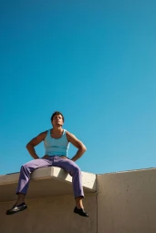 Model Sergio Modrego sits on a concrete ledge wearing a light blue tank top and purple trousers against a clear blue sky.