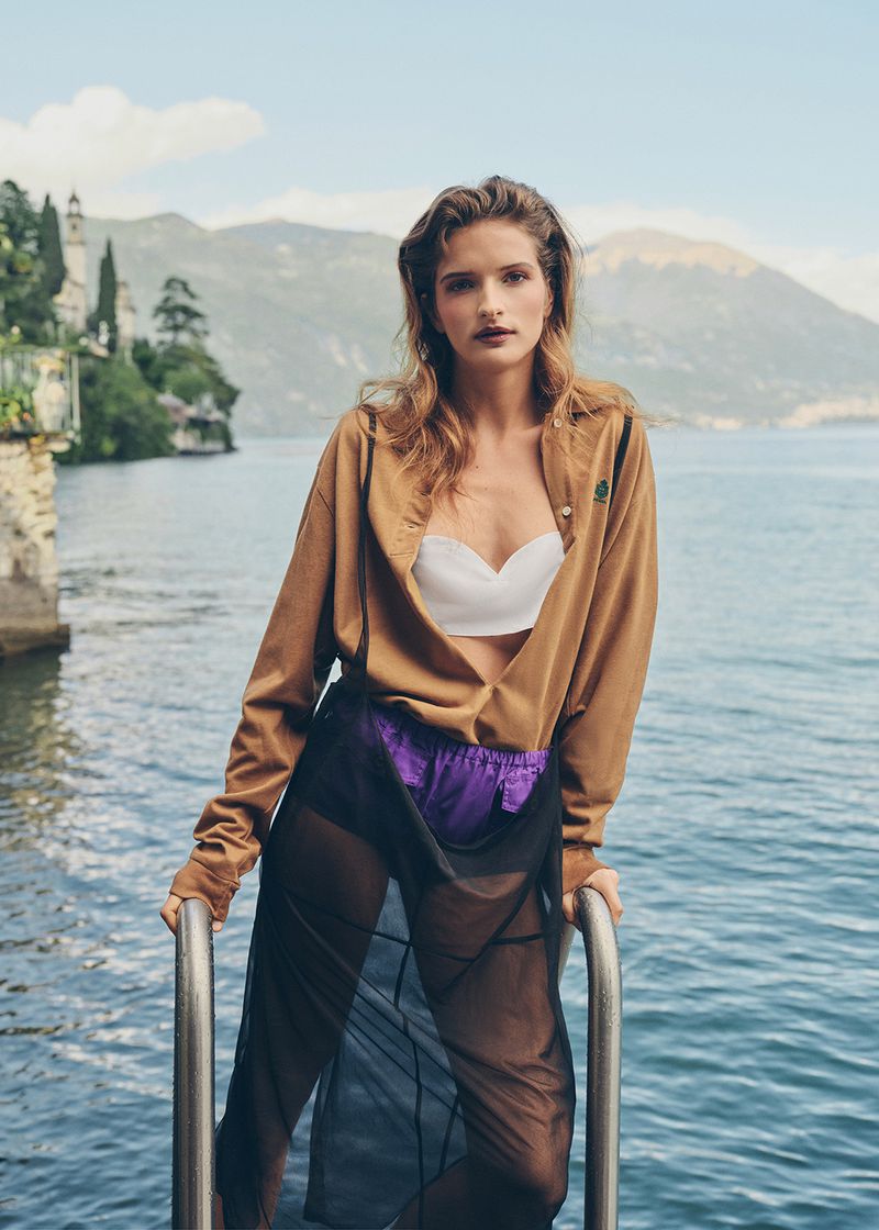 A woman in an unbuttoned tan shirt, white bralette, and sheer black skirt poses by a lake holding a metal dock ladder.